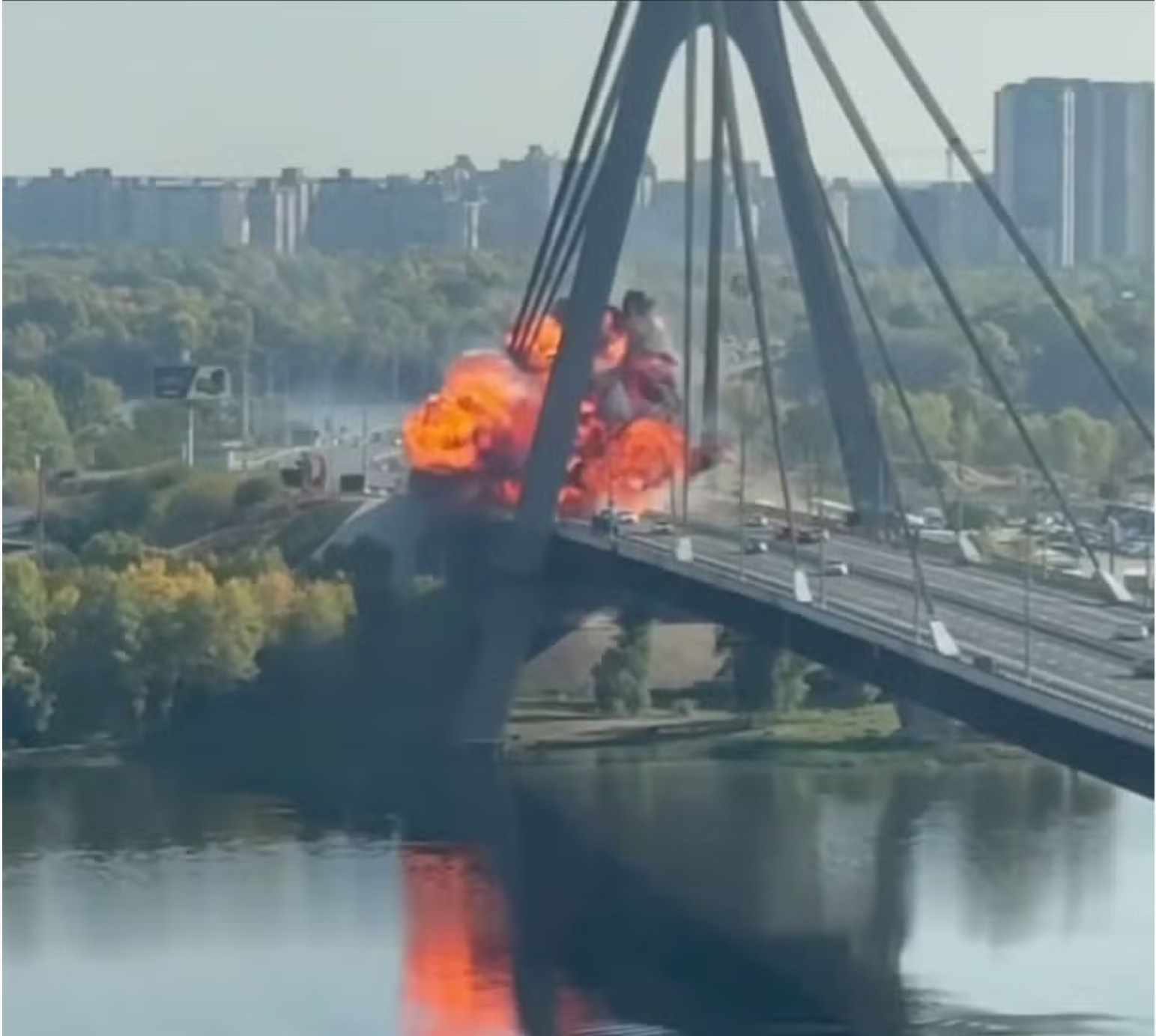 A fireball explosion at the tower of a cable-stayed bridge over a river, with cars on the deck