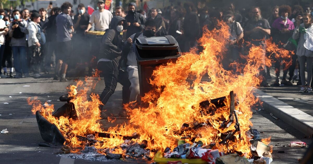 Masked protesters beside a burning pile of garbage and a wheelie bin in a street, with a crowd behind them